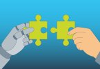 A robotic hand and a human hand are each holding a green puzzle piece that are meant to be put together.